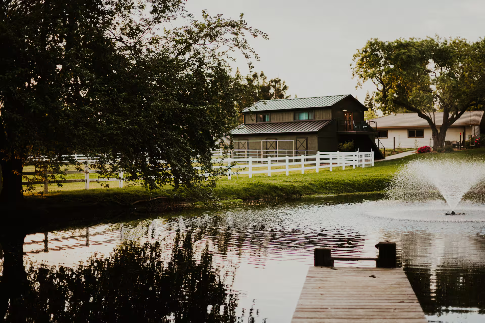 Dusk view of the barn with its green metal roof and balcony reflected in the pond, framed by mature trees, with a fountain on the right and a wooden dock extending into the foreground