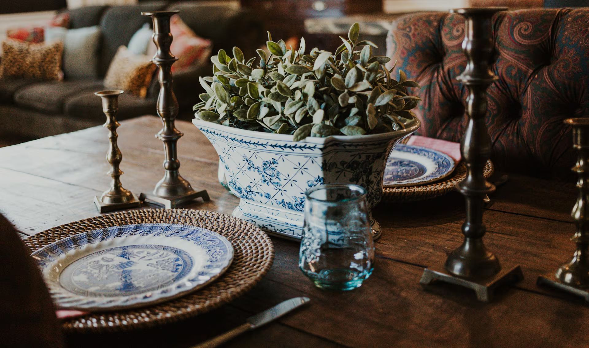 An editorial tablescape on a reclaimed wood farm table — blue willow-pattern dinner plate on a rattan charger, a blue-and-white Chinoiserie cachepot with fresh greenery, and tall brass candlesticks, with a paisley chesterfield sofa blurred in the background
